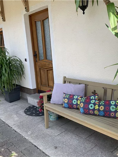 A cozy bench with colorful cushions stands in front of a front door. Surrounded by green plants and a paved path, the spot conveys an inviting atmosphere.