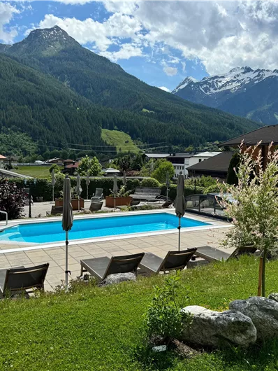 Ein schöner Pool in einer grünen Gartenlandschaft, umgeben von Bergen. Die Sonne scheint und der Himmel ist teilweise bewölkt.