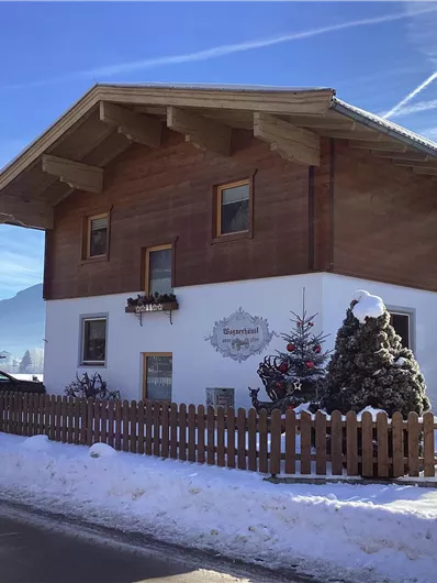 Ein charmantes Holzhaus im Schnee, umgeben von einer niedrigen Zaunreihe. Der klare Himmel und die Berge im Hintergrund verleihen der Szene eine idyllische Atmosphäre.