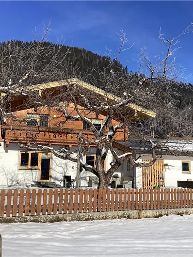 Ein traditionelles Holzhaus im Winter, umgeben von einer schneebedeckten Landschaft. Im Hintergrund sind verschneite Berge und ein blauer Himmel zu sehen.
