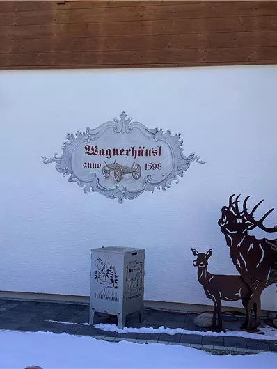 Ein traditionelles alpenländisches Gebäude mit einem dekorativen Schild. Draußen stehen eine Metallfigur eines Rentiers und ein geschmückter Weihnachtsbaum im Schnee.