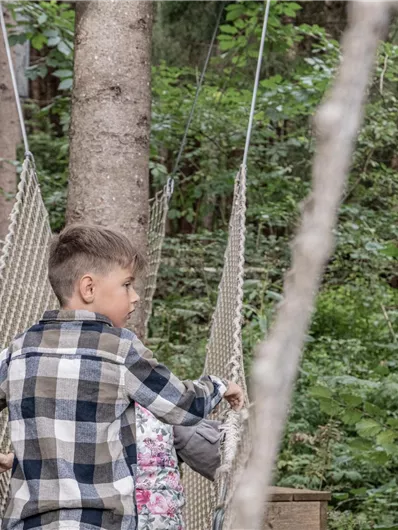Zwei Kinder stehen auf einer Seilbrücke im Wald. Um sie herum sind grüne Bäume und Pflanzen.