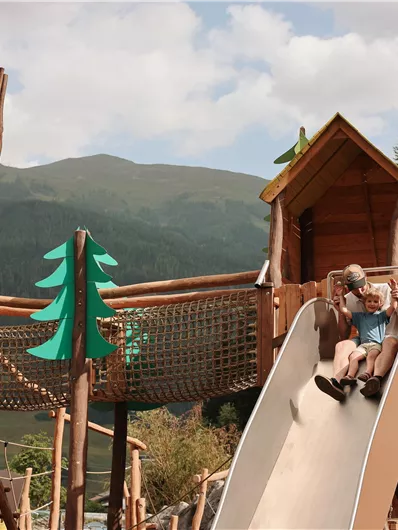 Ein Spielplatz mit einer Rutsche und Holzspielgeräten. Im Hintergrund sind grüne Berge und ein blauer Himmel zu sehen.