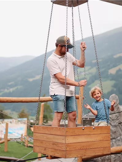 Ein Vater steht mit seinem Kind in einer Holzschaukel auf einem Spielplatz. Im Hintergrund sind grüne Berge und eine bewölkte Himmel zu sehen.