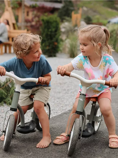 Zwei Kinder sitzen auf ihren Fahrrädern und schauen sich freundlich an. Im Hintergrund sind einige Spielgeräte und Natur zu sehen.