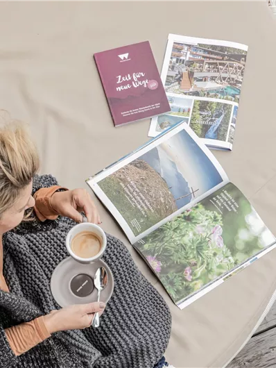 Eine Frau sitzt an einem Tisch und genießt eine Tasse Kaffee. Neben ihr liegen einige ansprechend gestaltete Broschüren und ein Buch.