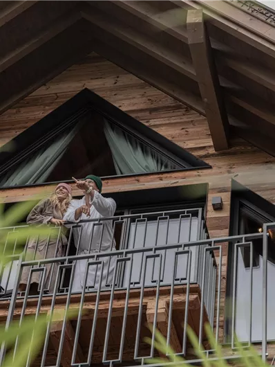 Een stel staat op een balkon van een houten huis en geniet van het moment. Op de achtergrond zijn groene planten te zien.
