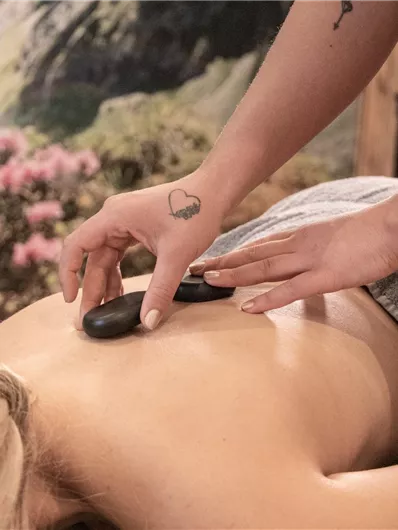 Eine Frau erhält eine Hot-Stone-Massage mit glatten Steinen auf ihrem Rücken. Im Hintergrund sind sanfte Farben und blühende Pflanzen zu sehen.