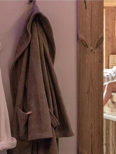 Ein gemütlicher Saunabereich mit zwei Bademänteln an der Wand. Im Hintergrund entspannen ein Paar in der Sauna.