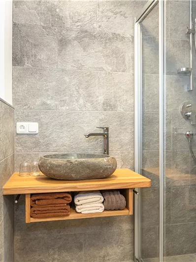 Een moderne badkamer met een douche en een stijlvol stenen wastafel. De houten onderbouw biedt ruimte voor netjes gevouwen handdoeken.