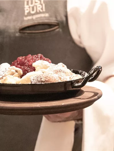Ein Kellner trägt eine kleine Pfanne mit einem Desserts. Das Gericht ist mit Puderzucker und roter Fruchtsoße garniert.
