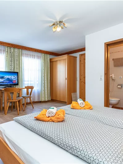 Een gezellige slaapkamer met een groot bed en dekens met een grijs patroon. Er is een televisie, een tafel en stoelen, evenals een zithoek.