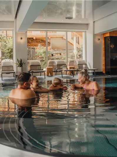 A family relaxes in the water of an indoor pool. The room is bright and modern with access to a beautiful garden area.