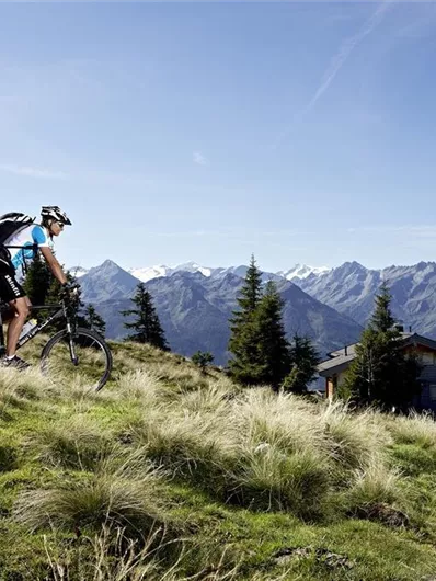 Twee mountainbikers rijden op een met gras bedekte heuvel. Op de achtergrond zijn besneeuwde bergen en een klein berghuis te zien.