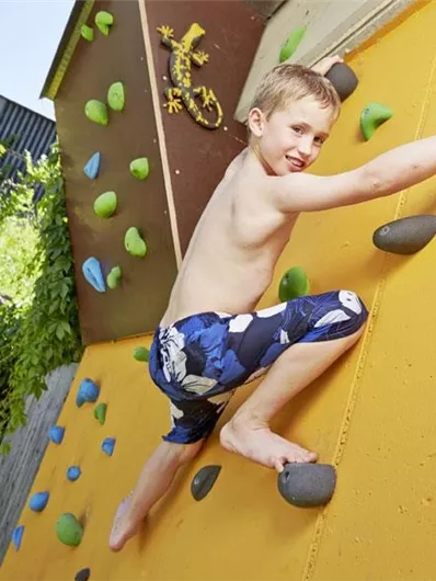 Een jongen klimt aan een kleurrijke klimwand in de buitenlucht. Hij draagt een zwembroek en toont veel energie en vreugde tijdens het klimmen.