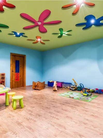 Ein bunter Spielraum für Kinder mit einem hellgrünen Deckel und farbenfrohen Blumenleuchten. Verschiedene Spielzeuge und Möbel sind verstreut, darunter ein Spieltisch und eine kleine Spielküche.