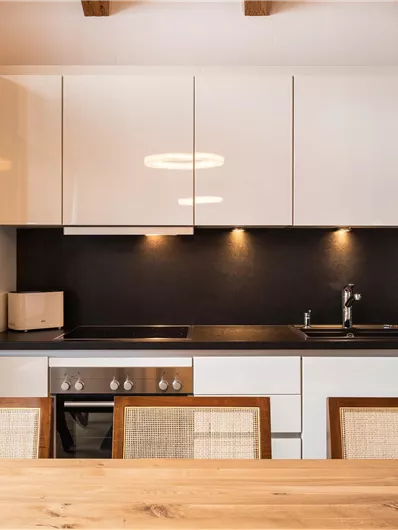 Eine moderne Küche mit weißen Möbeln und einer dunklen Arbeitsplatte. Im Vordergrund steht ein Holztisch mit Stühlen.