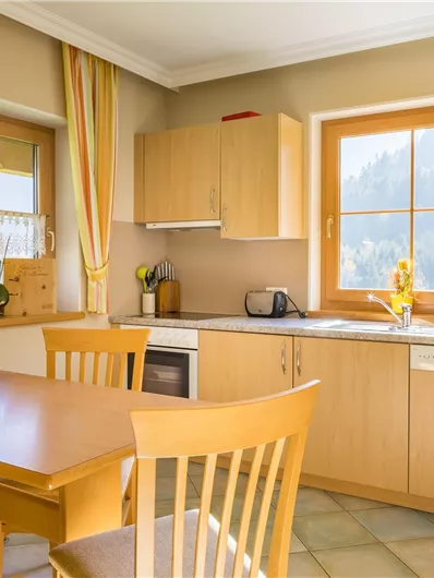 Een lichte en uitnodigende keuken met houten kasten en een kleine eethoek. Grote ramen laten veel daglicht binnen en bieden een mooi uitzicht op het landschap.