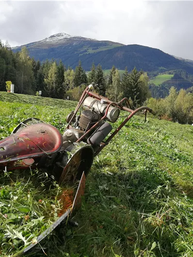 Ein altes Landmaschinenfahrzeug liegt auf einer Wiese in den Bergen. Im Hintergrund sind bewaldete Hügel und eine wolkige Landschaft zu sehen.