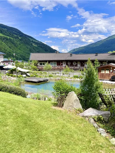 Eine malerische Landschaft mit einer Wiese, einem Teich und hübschen Blumen. Im Hintergrund sind Berge und Gebäude zu sehen.