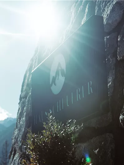 A sign with the inscription "Haus Holter" in the mountains. In the background, a sunny sky landscape and high mountains are visible.