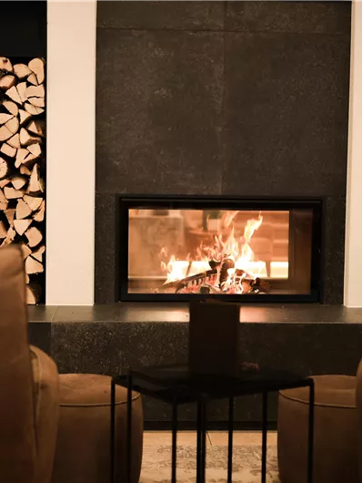 Ein gemütliches Wohnzimmer mit einem modernen Kamin, der ein warmes Feuer ausstrahlt. Neben dem Kamin stapeln sich Holzscheite und es stehen elegante Möbel im Raum.