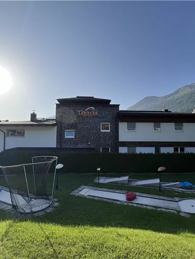 A modern building with several windows, surrounded by a green meadow. In the background, gentle hills and a radiant sky are visible.