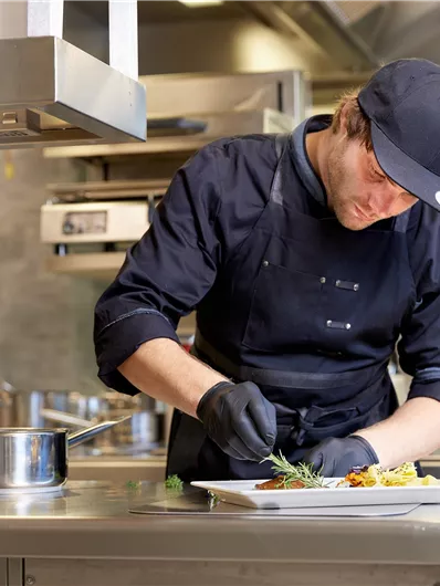 Een kok in een moderne keuken plateert een gerecht. Hij draagt een zwarte schort en handschoenen en concentreert zich op zijn werk.