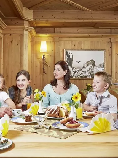 Een vrolijke familie zit aan een houten tafel en geniet van een ontbijt. De omgeving is gezellig met bloemen en traditionele inrichting.