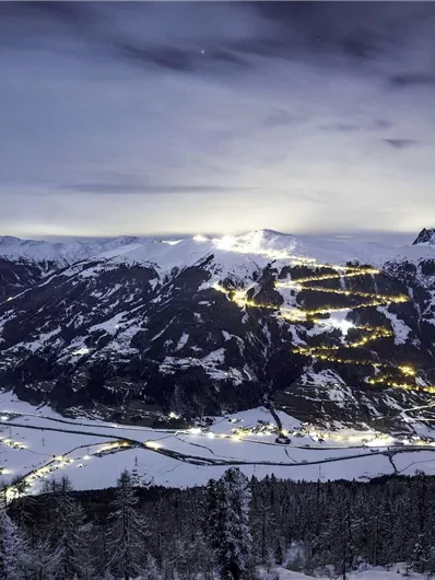 A snowy mountain landscape at night, illuminated by lights. The sky is clear with a gentle twilight.