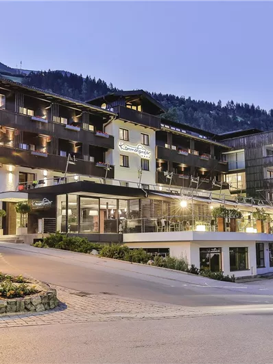 A modern hotel in the mountain landscape with stylish architecture. The scene is illuminated at dusk and shows an inviting atmosphere.