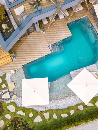 Ein modernes Schwimmbad mit Holzdeck und Sonnenschirmen in einem gepflegten Außenbereich. Die Umgebung ist mit Steinen und Grünflächen gestaltet.