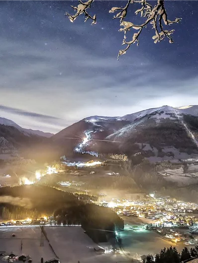 A wintry landscape at night with snow-covered mountains. The town in the valley glows warmly in the dark.