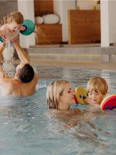 Eine Familie spielt im Wasser eines Schwimmbades. Zwei Kinder tragen Schwimmflügel und haben Spaß mit einem Erwachsenen.