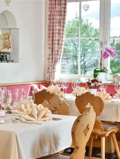 Een uitnodigende eetzaal met elegant gedekte tafels. De ramen laten veel licht binnen en de decoratie bestaat uit bloemen en frisse kleuren.