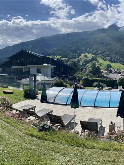 Ein schöner Garten mit einem Pool, umgeben von Bergen und einem blauen Himmel. Liegen und Sonnenschirme laden zum Entspannen ein.