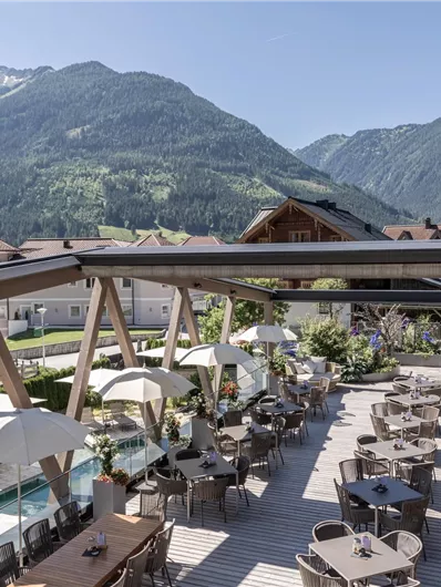 Ein modernes Restaurant im Freien mit Tischen und Sonnenschirmen. Der Blick umfasst die Berge und eine grüne Landschaft im Hintergrund.