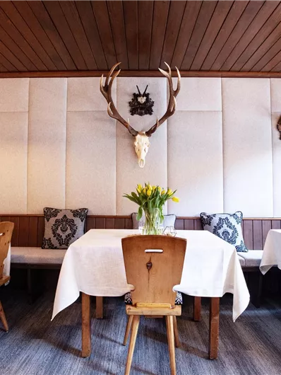 Ein gemütliches Restaurant mit Holzwänden und Tischen, die mit weißen Tischdecken gedeckt sind. An den Wänden sind Jagdtrophäen und eine Blumenvase auf dem Tisch ausgestellt.