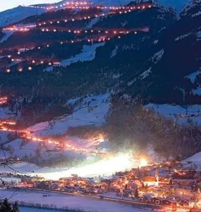 Een winterachtig berglandschap met met sneeuw bedekte hellingen en verlichte pistes. Op de voorgrond ligt een charmant dorp dat in warm licht straalt.