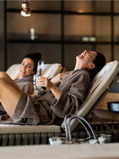 Two women are relaxing in a spa area and laughing while sitting on loungers in bathrobes. They are holding drinks in their hands and enjoying the relaxed atmosphere.