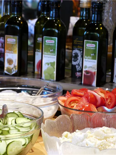 Een selectie van verse ingrediënten, waaronder komkommers, tomaten en kruiden. Op de achtergrond staan verschillende flessen olijfolie.