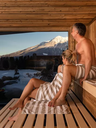 A couple relaxes in a sauna with wooden paneling. In the background, snow-covered mountains and a winter landscape can be seen.