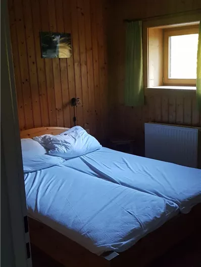 Ein einfaches Zimmer mit zwei Betten und Holzvertäfelung. Es gibt ein Fenster mit Vorhängen und eine kleine Wandmalerei.