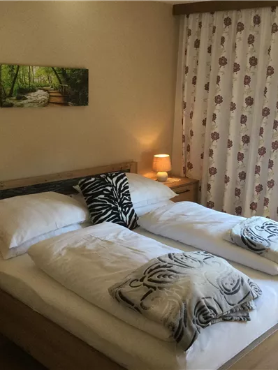 Ein gemütliches Schlafzimmer mit einem Doppelbett und sanfter Beleuchtung. Die Fenster sind mit Vorhängen dekoriert, und ein Bild von einer grünen Landschaft hängt an der Wand.