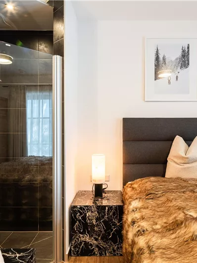 Ein modernes Schlafzimmer mit einem gemütlichen Bett und weichen Kissen. Im Hintergrund sind ein Badezimmerbereich und stilvolle Wandbilder zu sehen.