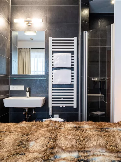 Ein modernes Badezimmer mit dunklen Fliesen und einer Dusche. Im Vordergrund ist ein komfortables Bett mit einer schönen Decke zu sehen.