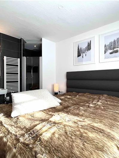 Ein modernes Schlafzimmer mit einem großen, gemütlichen Bett und einem Fellüberwurf. Im Hintergrund sind ein Badezimmerbereich und einige hübsche Wandbilder zu sehen.