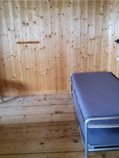 Ein einfaches Zimmer mit Holzverkleidung. Es enthält ein Bett und einen Stuhl, die einen minimalistischen Look haben.