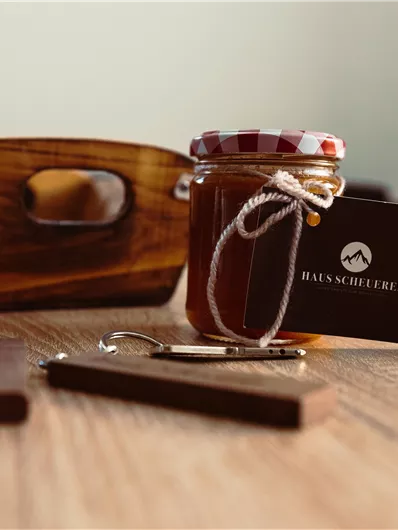 A glass of honey with a checked lid and a small card lies on a wooden table. Next to the glass are some wooden keychains and a small basket.