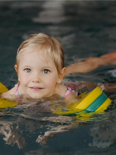 Ein kleines Kind schwimmt im Wasser mit Schwimmnudeln. Im Hintergrund lächelt eine Frau und beobachtet es freundlich.